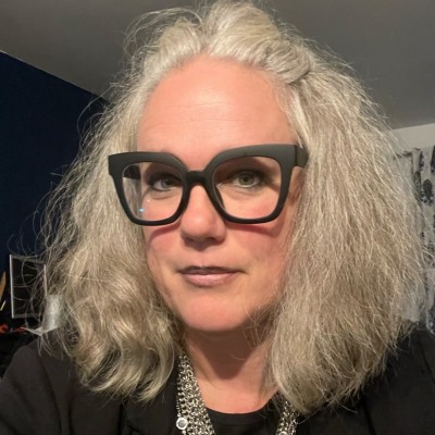 Portrait of a woman with shoulder-length gray hair and large black glasses, looking at the camera.