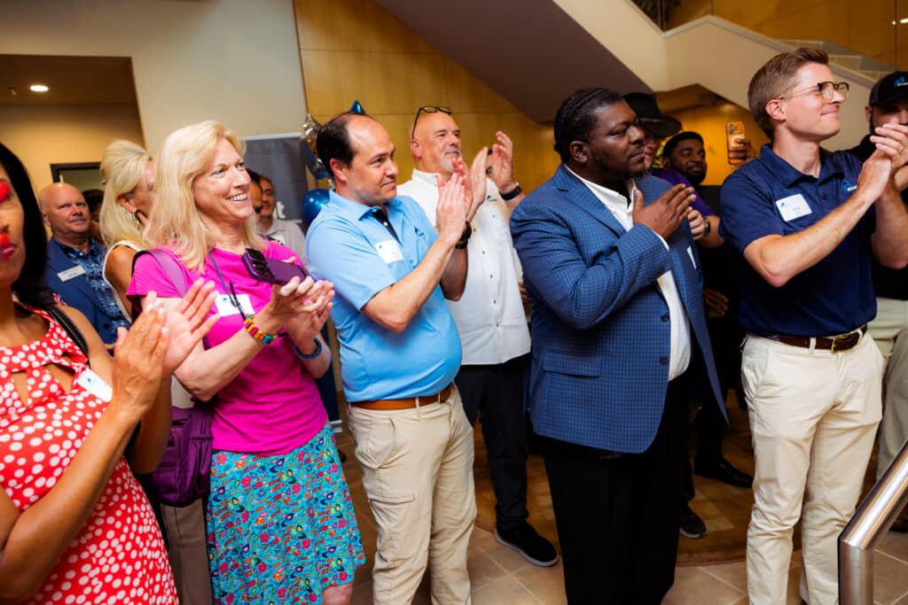 Group of people indoors, standing in a line, clapping and smiling at an event, some wearing name badges and colorful outfits.