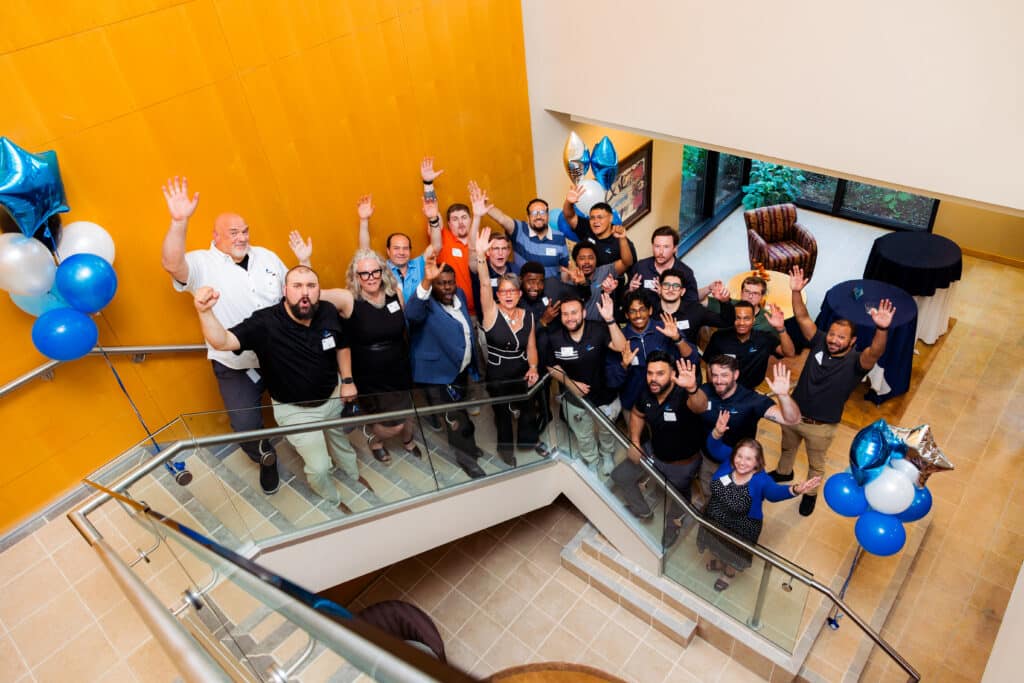 Group of diverse colleagues waving and smiling at camera in a bright atrium with orange wall and blue balloons, celebrating an event.