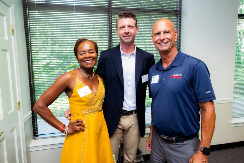 Three smiling adults posing together indoors near a window with blinds; woman in yellow dress, two men in business and polo shirts wearing name badges.