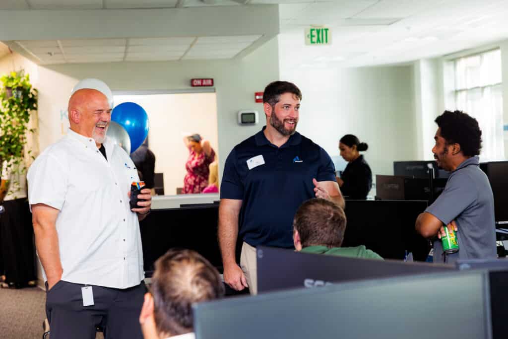 Group of colleagues chatting in a bright office; man in white shirt smiles on the left while another in a navy polo speaks center.