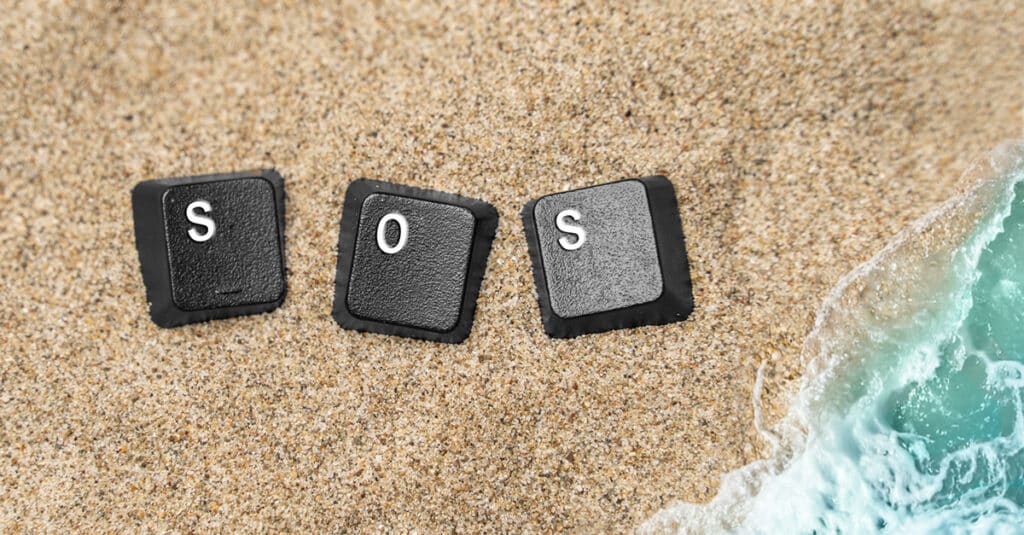 Three black keyboard keys spelling SOS lie on sandy beach with the turquoise sea nearby.