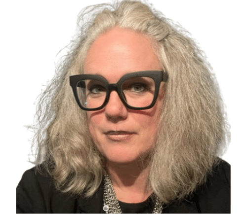 Person with shoulder‑length gray hair and oversized black glasses looking at the camera in a dark outfit.