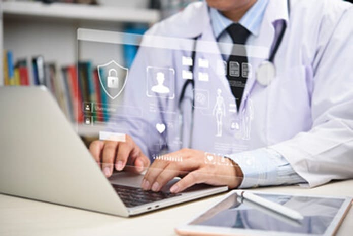 Doctor in a white coat typing on a laptop with translucent holographic medical icons overlaying the screen.