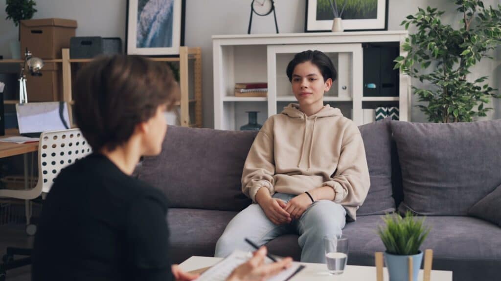 Therapist or interviewer speaks with a young person sitting on a gray sofa in a cozy living-room therapy setting.