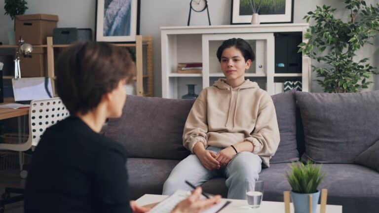 Therapist or interviewer speaks with a young person sitting on a gray sofa in a cozy living-room therapy setting.