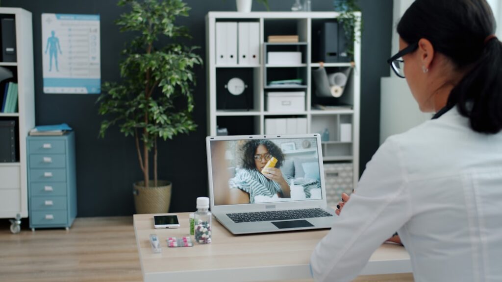 Telemedicine scene: a healthcare professional on a video call with a patient shown on the laptop screen, with pills and medication on the desk nearby.